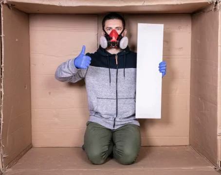 Man in a protective mask is sitting in a cardboard box, holding an empty boar Stock Photos