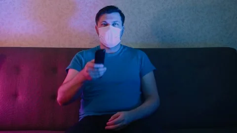 A man in a protective mask sitting on the couch carefully watches television Stock Footage 130973903