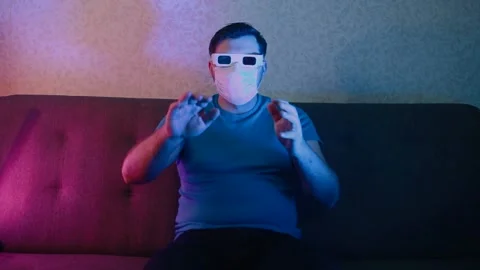 A man in a protective mask is sitting on a sofa wearing stereo glasses, watching 스톡 동영상 131736989