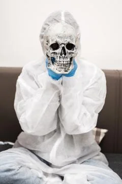 Man in protective white translucent suit, blue rubber gloves, medical mask sits Stock Photos