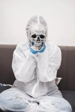 Man in protective white translucent suit, blue rubber gloves, medical mask sits Stock Photos