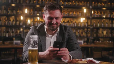 The man is at the pub, drinking beer and surfing on his phone. A handsome man is Video stock 278470831