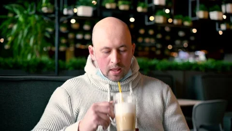 Man pulled off face mask for drinkig cappuccino on terrace. Man in cafe outdoors Stock Footage 149000364