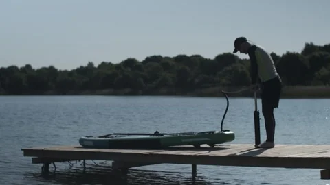 Man pumping stand up paddle board standing on wooden bridge on lake shore. SUP Stock Footage 243428328