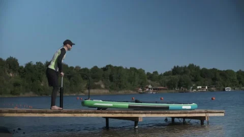 Man pumping stand up paddle board standing on wooden bridge on lake shore. SUP Stock Footage 243428651