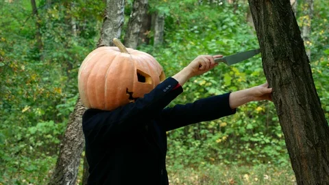 A man with a pumpkin on his head. He pulls out a knife from a tree. Halloween. Video stock 116495373