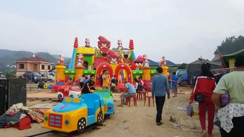 Man pushes circular plastic train, kids on inflatable bouncer, funfair, Sam Neua Vídeos de archivo 76927581