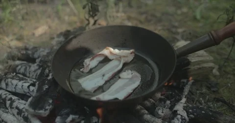 Man puts bacon into frying pan on campfire Stock Footage 76703458