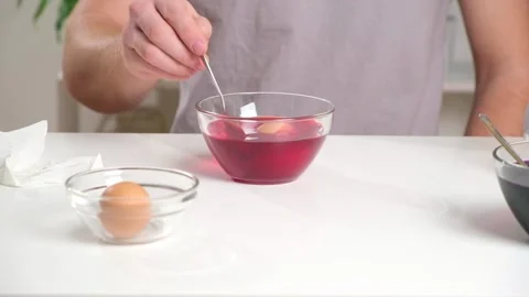 A man puts an egg on a spoon into a glass cup with a dye. Dyeing eggs with Stock Footage 236660074