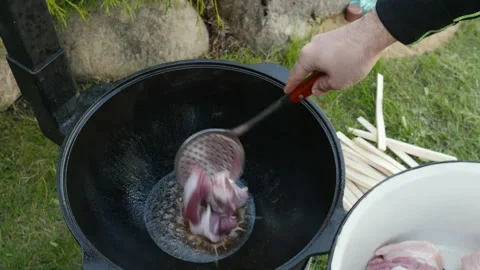A man puts meat at cauldron. Cooking process, frying pork meat open fire.  Vidéo 131542490