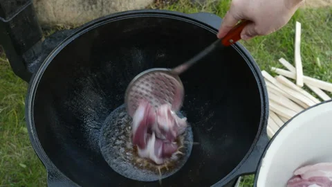 A man puts meat at cauldron. Cooking process, frying pork meat on open fire.  Stock Footage 131544435