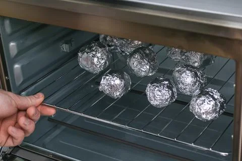 A man puts raw potatoes wrapped in foil on wire rack in the oven. Stock Photos