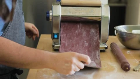 Man puts sheet of beet purple pasta dough through a pasta rolling machine Stock Footage 199153012