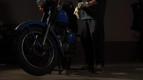 Man putting on gloves, preparing to work on a classic blue motorcycle in a Stock Footage 342760770
