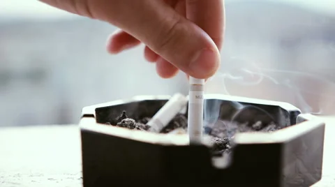 Man putting out a smoking cigarette in an ashtray with buildings in background Stock Footage