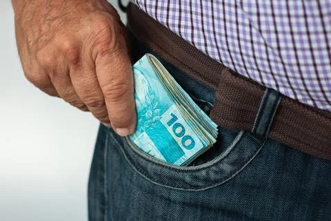 Man putting a stack of 100 Brazilian real banknotes into his jeans pocket Stock Photos