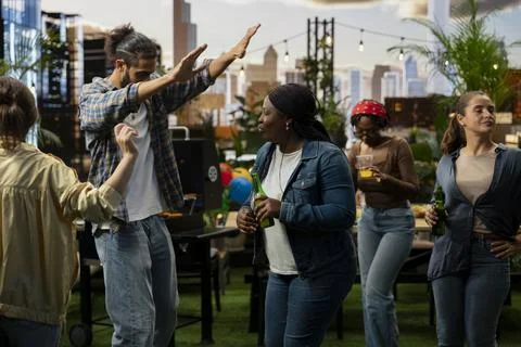 Man raising arms while dancing beside woman holding drink at rooftop BBQ party Stock Photos