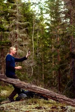 Man raising phone in forest while resting on fallen tree Stock Photos