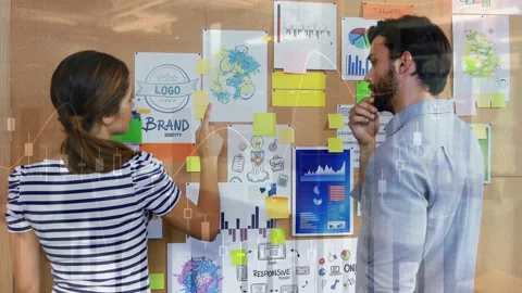 Man reaching sticky note on corkboard while woman arranging brand charts for Stock Footage 332625464