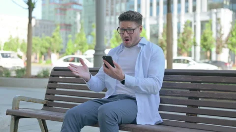 Man Reacting to Loss on Smartphone while Sitting Outdoor on Bench Stock Footage 197738129