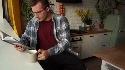 A man reading a book and drinking coffee while sitting in the kitchen Stock Footage 157885871