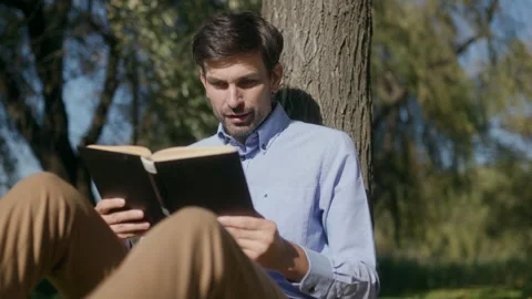 Man reading book and looking at camera leaning on tree in park, cinematic Stock Footage 332298937