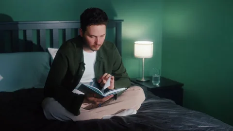 Man reading book in bed under warm lamp light Vídeos de archivo 319544412