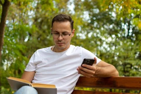 A man is reading a book on a bench while checking his phone Stock Photos