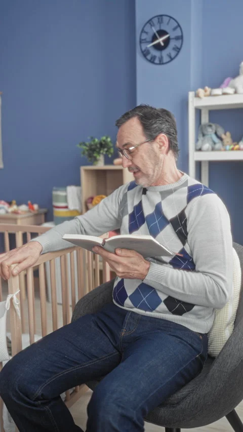 Man reading book, holding page and touching crib rail beside wall clock in .. Stock Footage 337443023