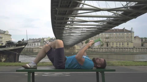 Man is reading book, lying on the bench against the river. Vídeo Stock 65188204