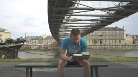 Man is reading book, sitting on bench against the river. 库存影片 65188108