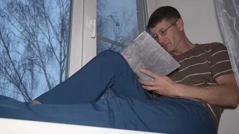 A man reading a book sitting on a windowsill. Stock Footage 97784410