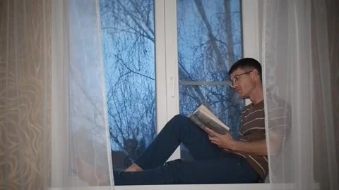 A man reading a book sitting on a windowsill. Stock Footage 97784425