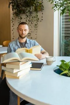 Man reading a book at a table with stacks of books and a cup in a modern sp.. Фото