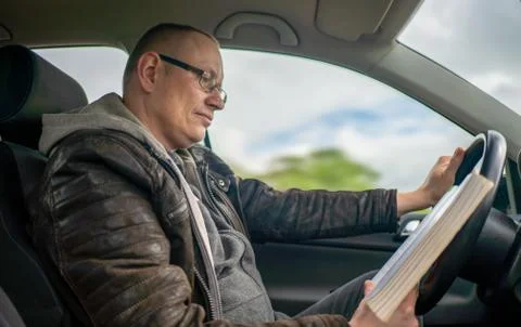 Man is reading a book while driving a car and is distracted Stock Photos