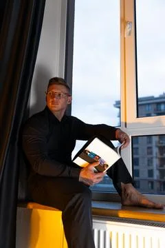 Man reading a book on a windowsill while enjoying a quiet evening in a modern Stock Photos