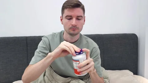 Man reading label on white plastic supplement bottle while sitting in bed Stock Footage 284041961