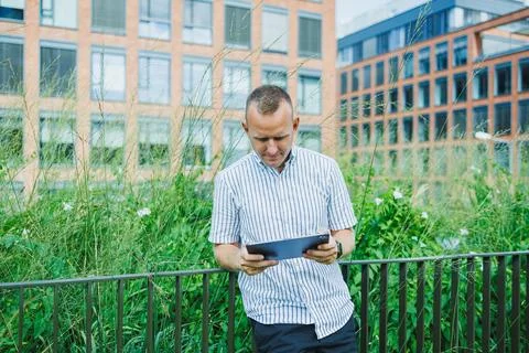 Man reading a tablet while leaning on a fence near green plants and modern .. Stock Photos