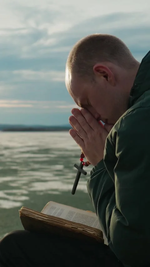 A man reads the Bible, praying to the Lord God on the shore of a lake. Stock Footage 344021167