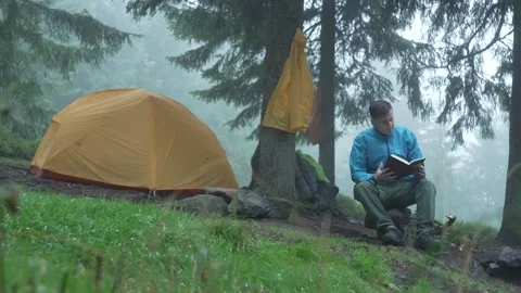 A man reads a book in the forest in rainy weather, camping near a tent. Fog Stock Footage 217395343