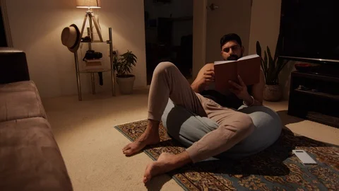 Man Reads Book in Living Room at Night, Phone lays forgotten. Wide Shot Stock Footage
