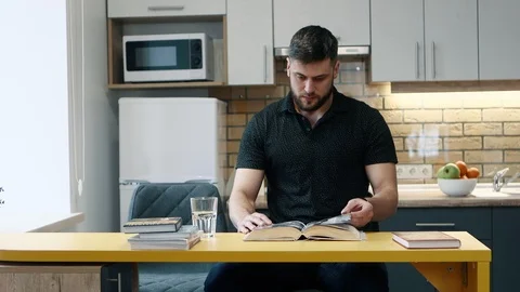 Man reads a book sitting in the kitchen at home and drinks water. Stock Footage 103855965