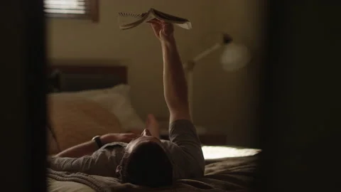 Man reads a book while holding it over his face while laying on a bed 스톡 동영상 134280234