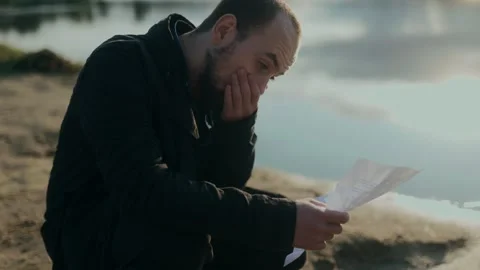 Man reads emotional letter while grieving alone by the calm lake Stock Footage 313606448