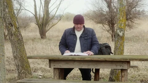 A man reads a thick book sitting at a wooden table between the trees Vídeos de archivo 171649985