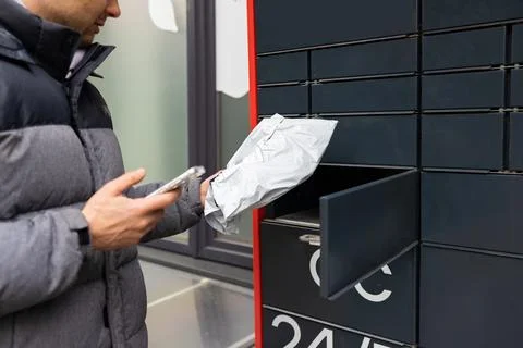 Man receiving parcel from automatic post box using smartphone outdoors. Stock Photos