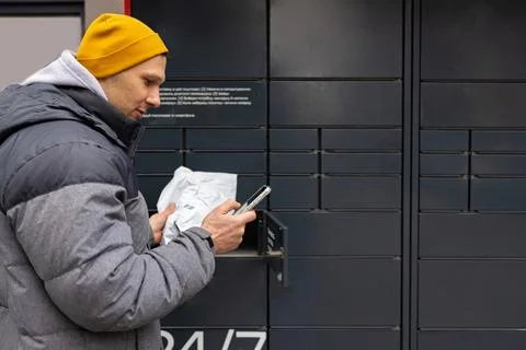 Man receiving parcel from automatic post box using smartphone outdoors. Photos