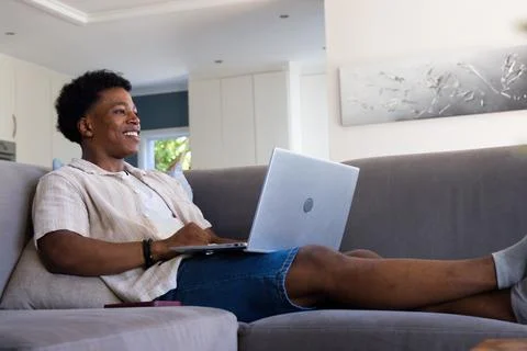 Man reclining on grey sectional sofa at home using silver laptop with smartphone Stock Photos