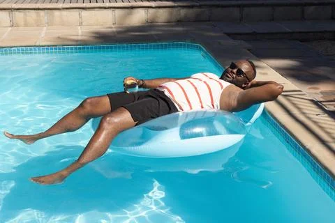 Man reclining on pale blue float at home pool edge, holding drink, wearing Stock Photos