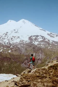 Man with red backpack stand on rocky mountain and look at snowy peak of Elbrus. Stock Photos